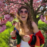 A person in a colorful sweater and brown tanktop poses for a photo beneath a tree of flowering pink photos