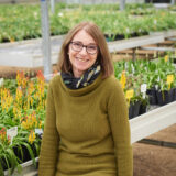 A person in a green sweater and black scarf poses for a photo in a greenhouse of plants
