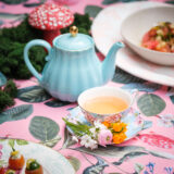 a light blue teapot sits on a pink printed tablecloth with a floral teacup in front filled with tea