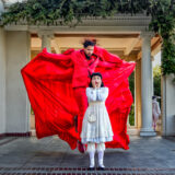 A person in vivid red clothing surrounds another person wearing a white dress, as if menacing them