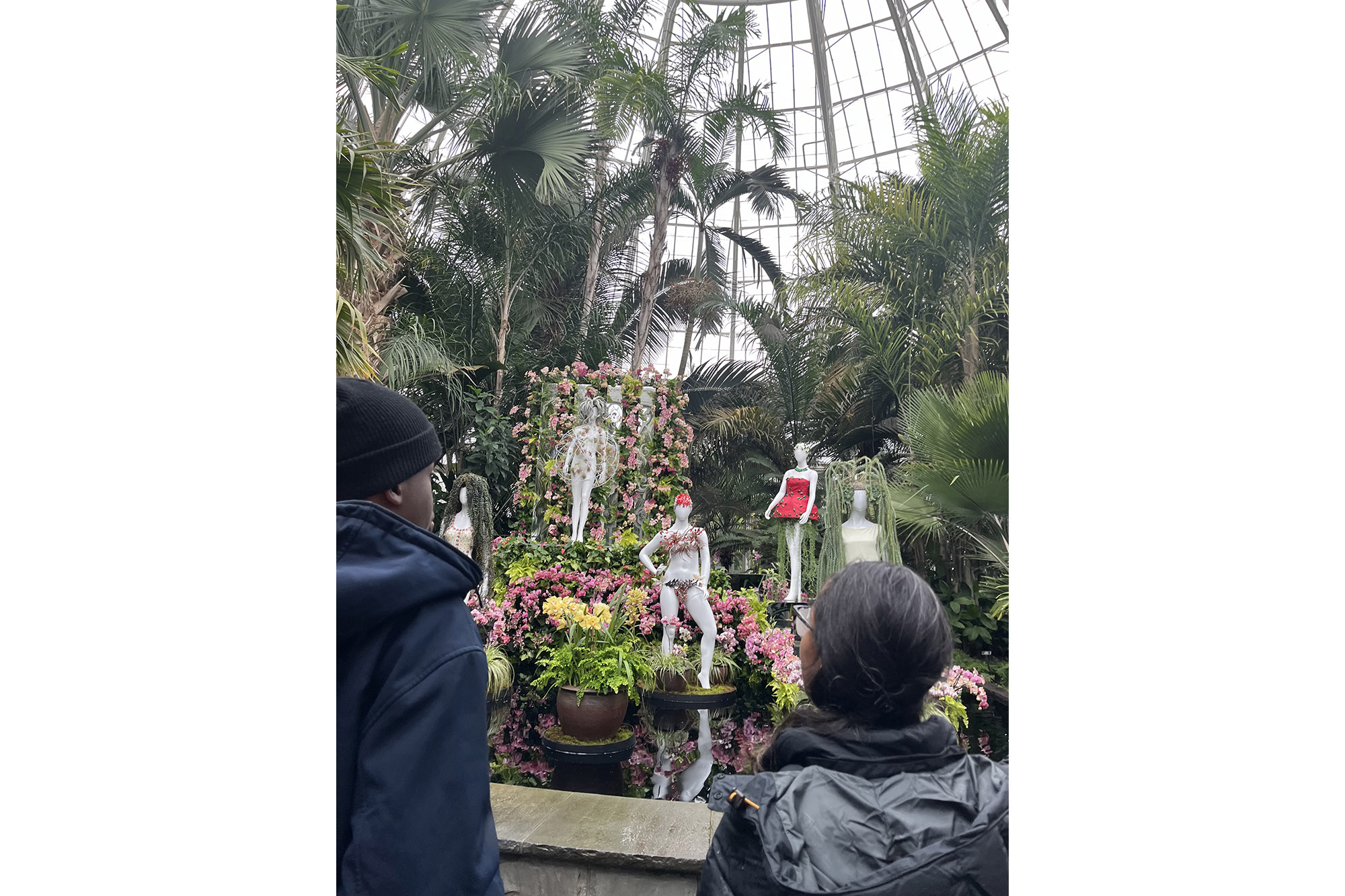 Students in winter clothes explore a lush green conservatory full of mannequins dressed with living plants
