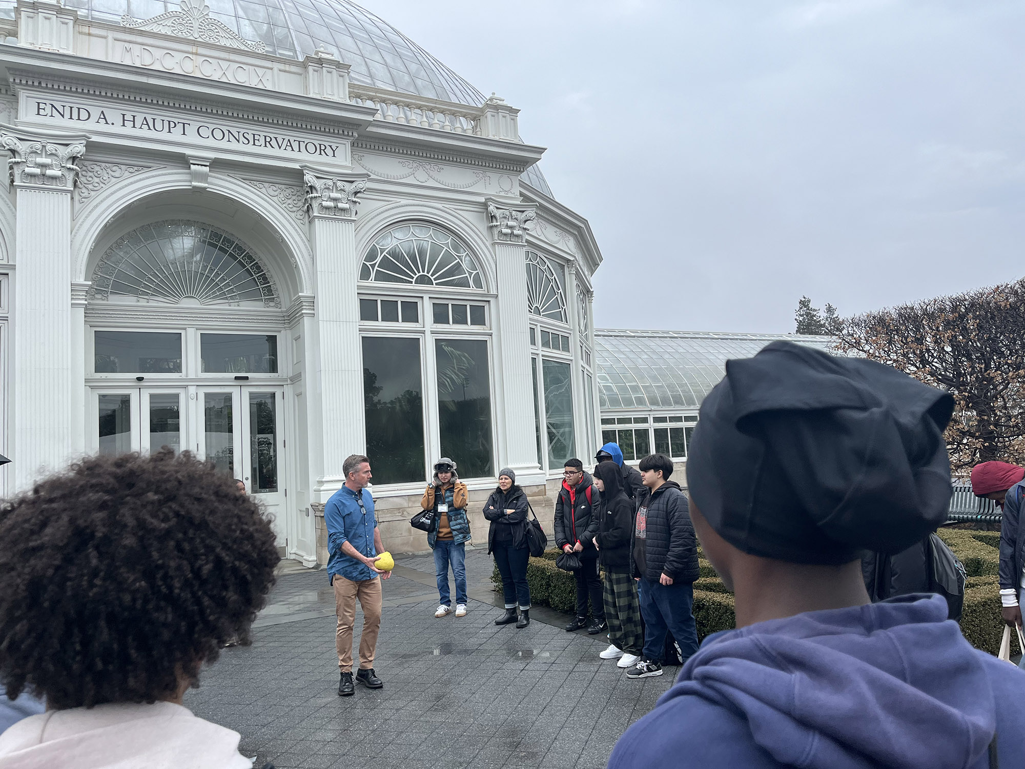 A person shows a large yellow citrus fruit to a group of high school students outdoors on a rainy day