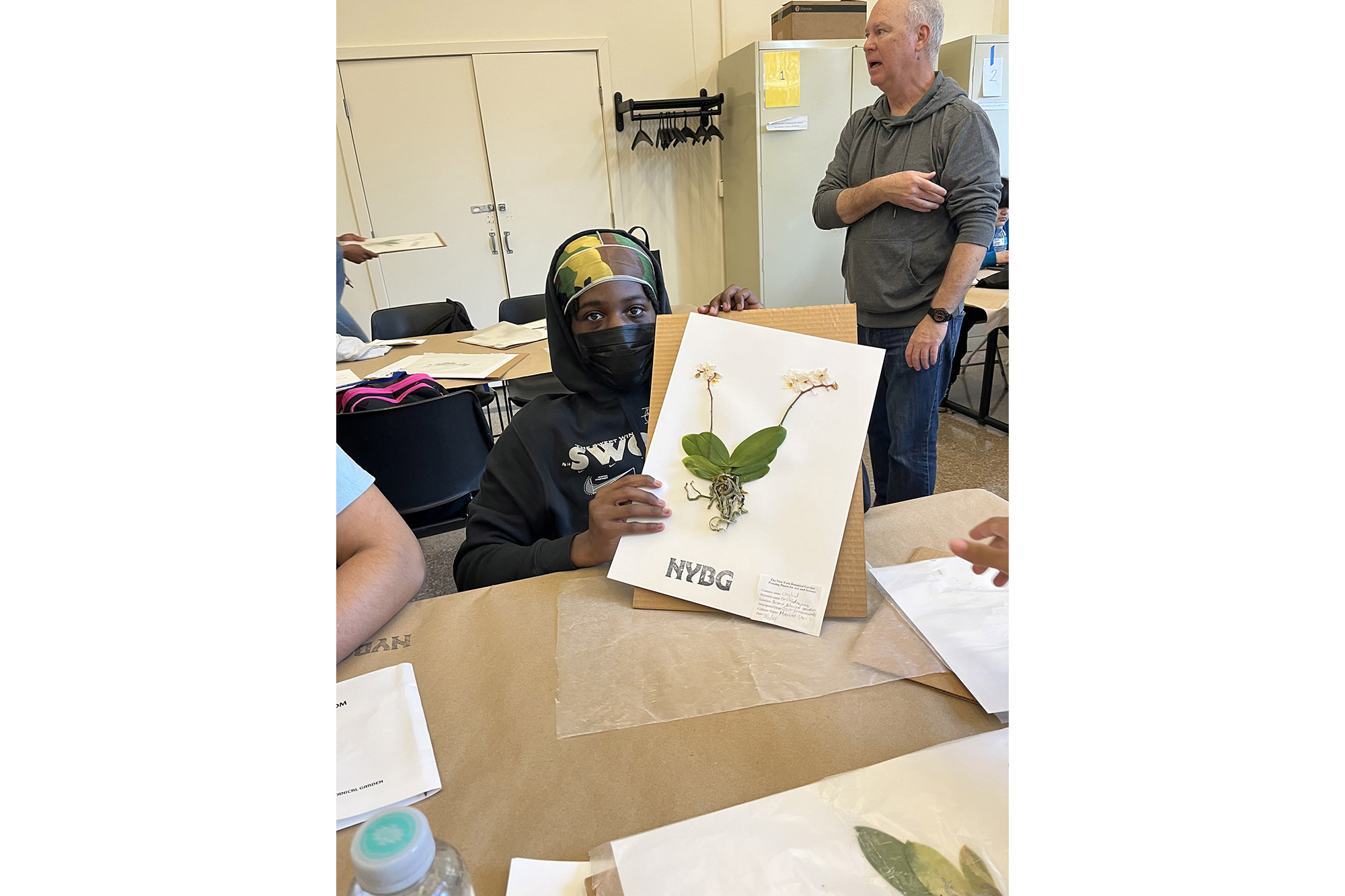 A student wearing a black mask holds up an herbarium specimen they made