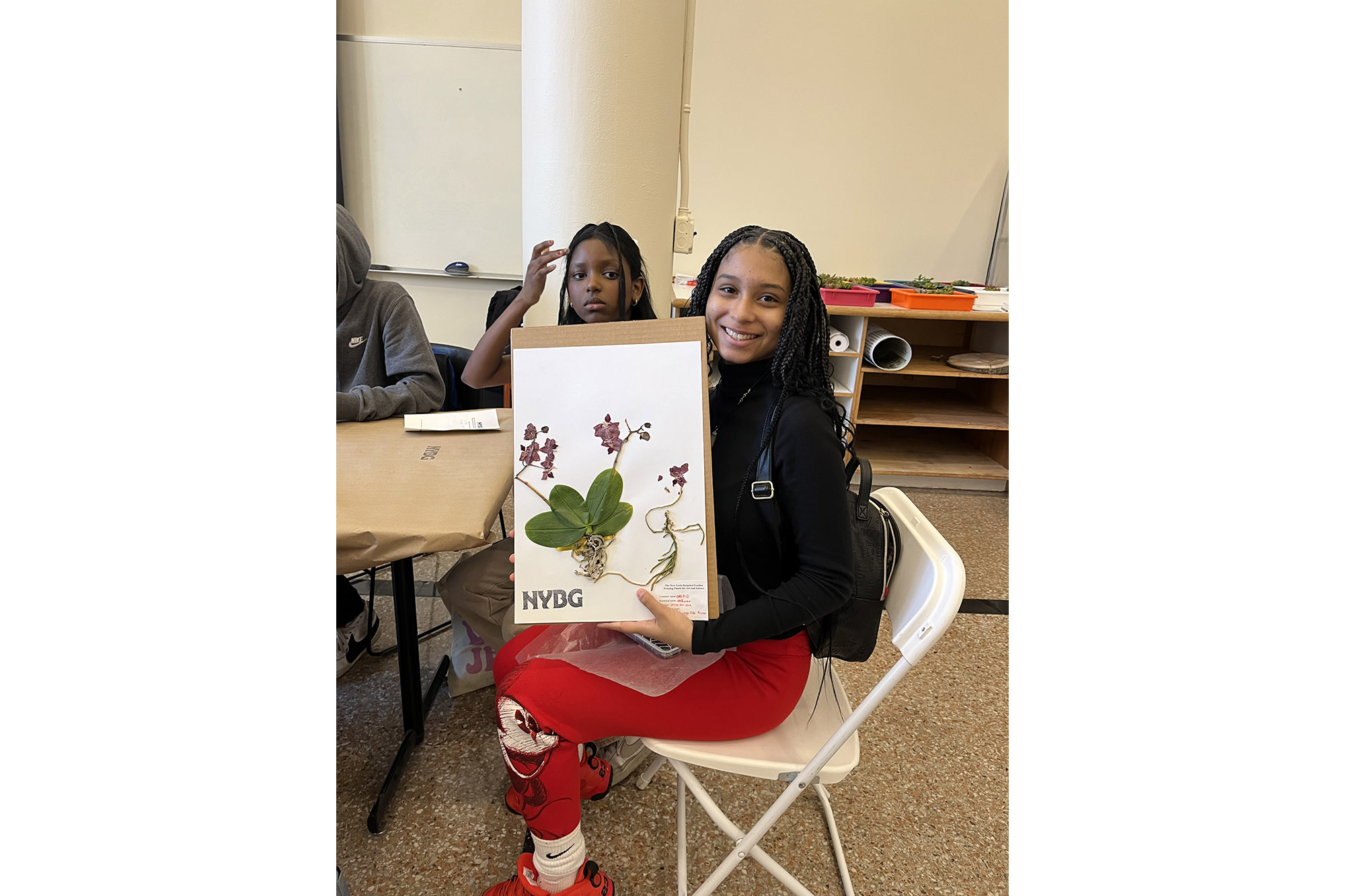 A student in red pants and a black shirt holds up an herbarium specimen they made