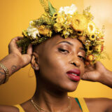 A person in a yellow shirt, wearing a crown of bright yellow flowers, poses for a photo in front of a yellow wall