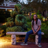 A person in blue jeans and a button up shirt sits for a photo next to an elaborately pruned green topiary