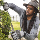 A person in a gray bucket hat with long dark hair uses clippers to prune a green topiary