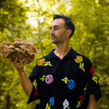 A person in a black shirt covered in colorful flowers holds a large cluster of mushrooms
