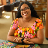 A person with long dark hair, wearing a colorful floral blouse, poses for a photo in a sunny library