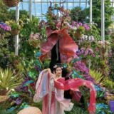 A person in a flowing, colorful dress poses in front of a vivid floral display