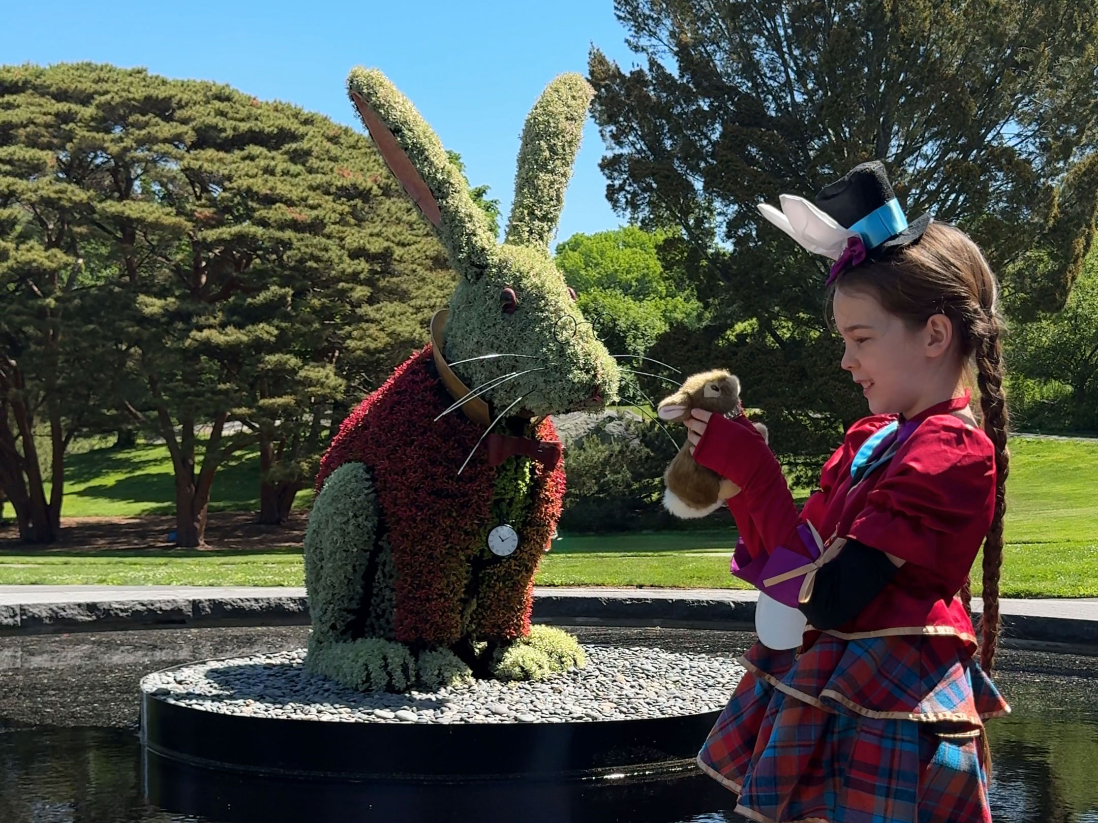A child in a colorful outfit poses for a photo near a giant rabbit topiary made of plants