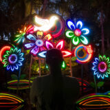 A person photographs a colorful evening display of flower sculptures lit up in a rainbow of hues