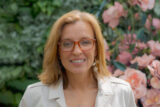 A person in red glasses and a white jacket poses for a photo in front of green leaves and pink flowers