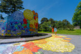 Two people in spring dresses walk around a monumental art installation of yellow, orange, and purple flowers