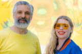Two people pose for a photo in front of a bright yellow wall of painted flowers