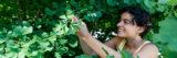 A person with dark curly hair and a yellow tank top smiles and examines a leaf on a green tree with a magnifying glass.