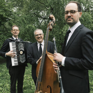 Three people in black suits pose with instruments outdoors