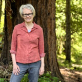 A person in a pink shirt and jeans poses for a photo in front of an enormous tree trunk