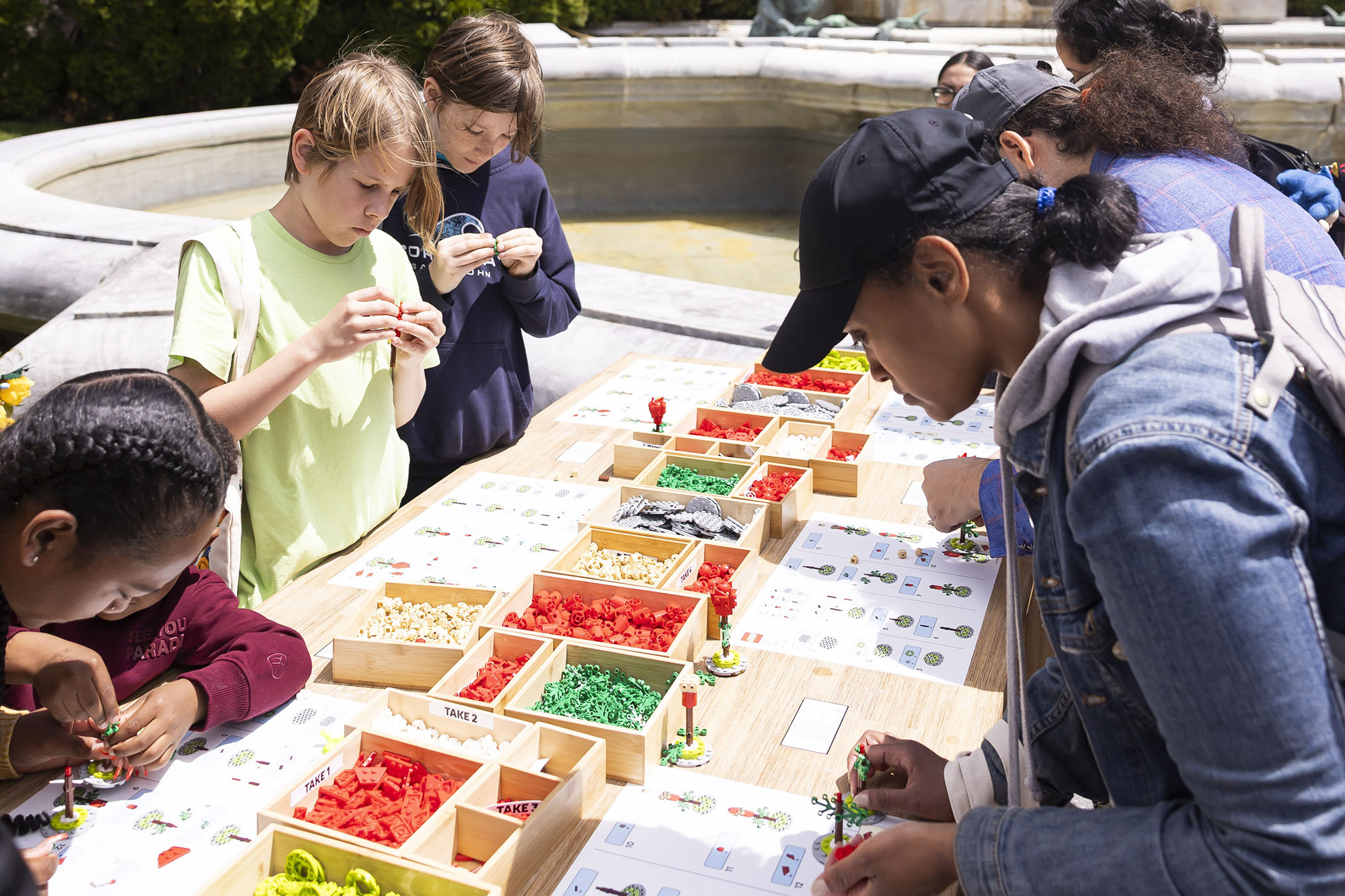 People use small, colorful parts to create LEGO flowers outdoors on a sunny day
