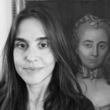 A person with long dark hair poses for a black and white photo with a painting in the background