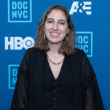 A person in a black shirt and necklace poses for a photo in front of a blue step-and-repeat wall
