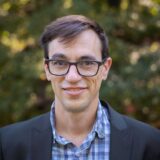 Person with brown hair, glasses, blazer, and blue checkered shirt looking straight ahead