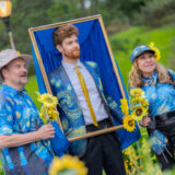 a person within a frame in The Starry Night-inspired blazer poses with two people holding sunflowers and in tee shirts that are inspired by The Starry Night