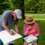 two people in hats sit outdoors looking at sketchbooks