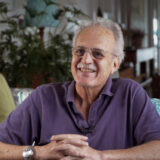 A person in a purple shirt and glasses sits while posing for a photo