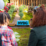 A parent and child watch a model train on a track, surrounded by green vegetation