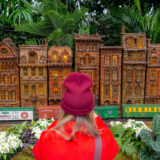 A person in a red beanie and sweater photographs a display of small brown building replicas, as a model train passes by