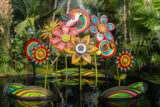 An arrangement of large flower sculptures in pink and yellow and blue sit in a pool of water, surrounded by green vegetation