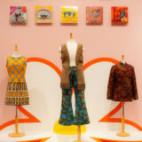 An arrangement of colorful outfits on dress forms under a row of small square paintings