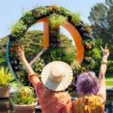 Two people throw up peace signs in front of a large botanical peace sign covered in flowers