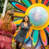 A person in a black dress poses for a photo in front of a large rainbow-colored flower sculpture