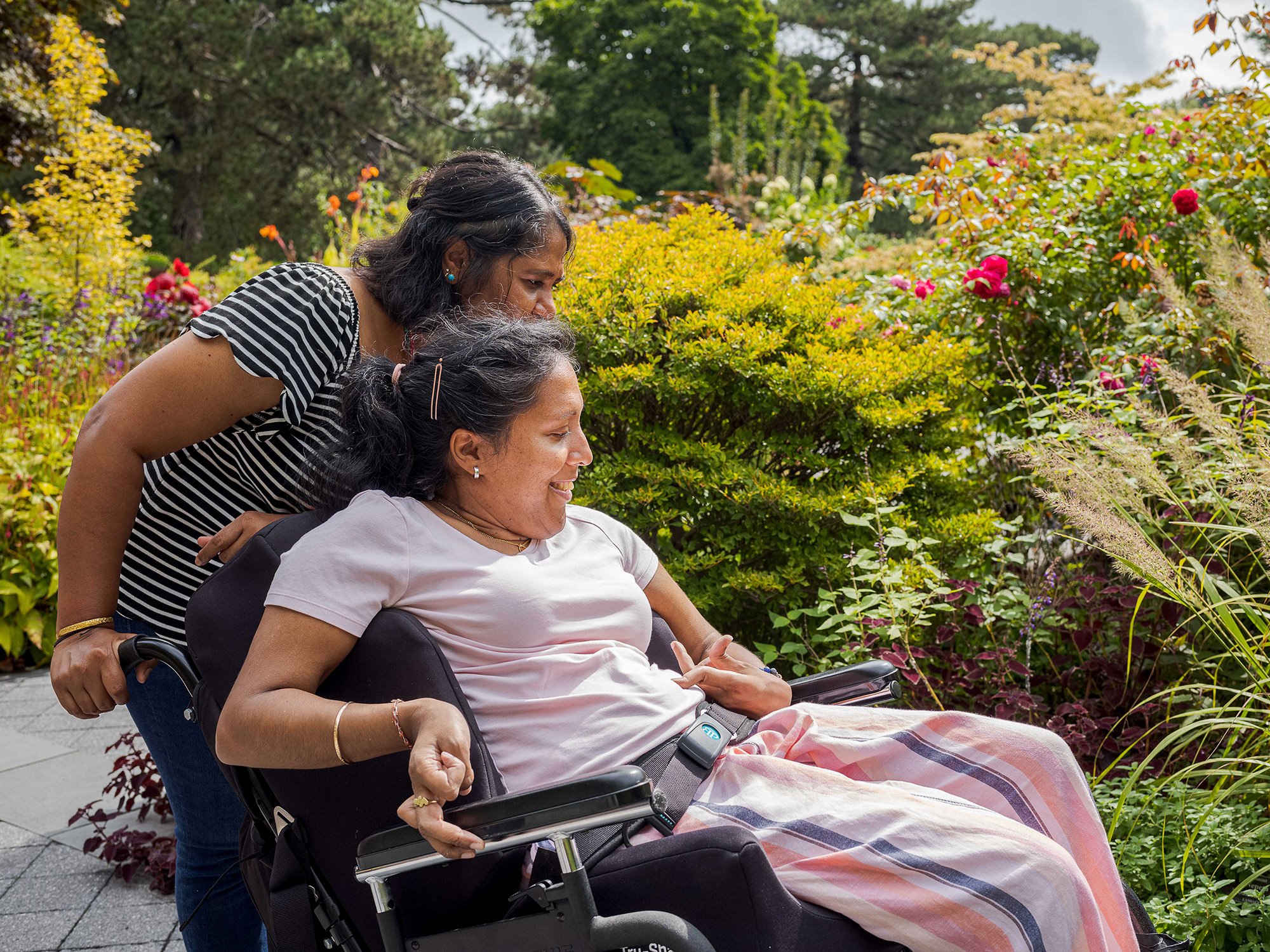 One person pushes another using a wheelchair through a sunny garden setting