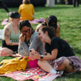 Two people sit on a picnic blanket, laughing over something on a phone