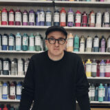 A person in a hat, glasses, and black shirt poses for a photo in front of a wall of artist paints