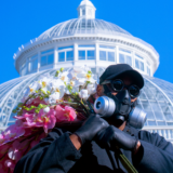A person in a black outfit holding bundles of pink and white flowers stands before a bright white conservatory dome, with a clear blue sky above