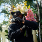 A person in a mask and black outfit holds bundles of pink and white flowers in a sunny conservatory setting