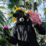 A person in a mask and black outfit observes a pink flower while carrying bundles of pink and yellow flowers on their back