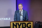 A person in a blue suit speaks at a lectern labeled 
