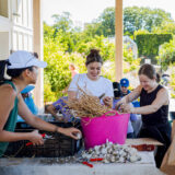 Three people place garden vegetables in a pink bin