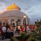 A large party of people enjoy an evening event beneath the dome of a conservatory