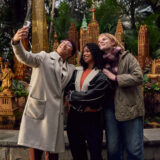 Three people take a selfie among a warmly lit holiday display of miniature buildings