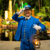 A person in a blue train conductor's outfit poses for a photo in front of a brightly lit holiday display
