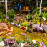 A colorful toy train travels through a lushly planted miniature cityscape