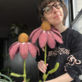 A person wearing glasses and a black sweatshirt poses with two pink and yellow crocheted flowers