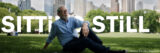 A person lounging on a park lawn in a city, with the words "Sitting Still" behind them
