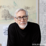 A person in a black sweater and glasses sitting for a photo in front of a number of landscape illustrations